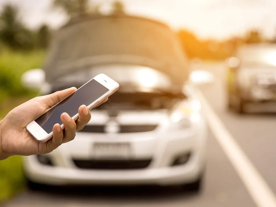Driver holding a phone in front of a broken-down car on a road
